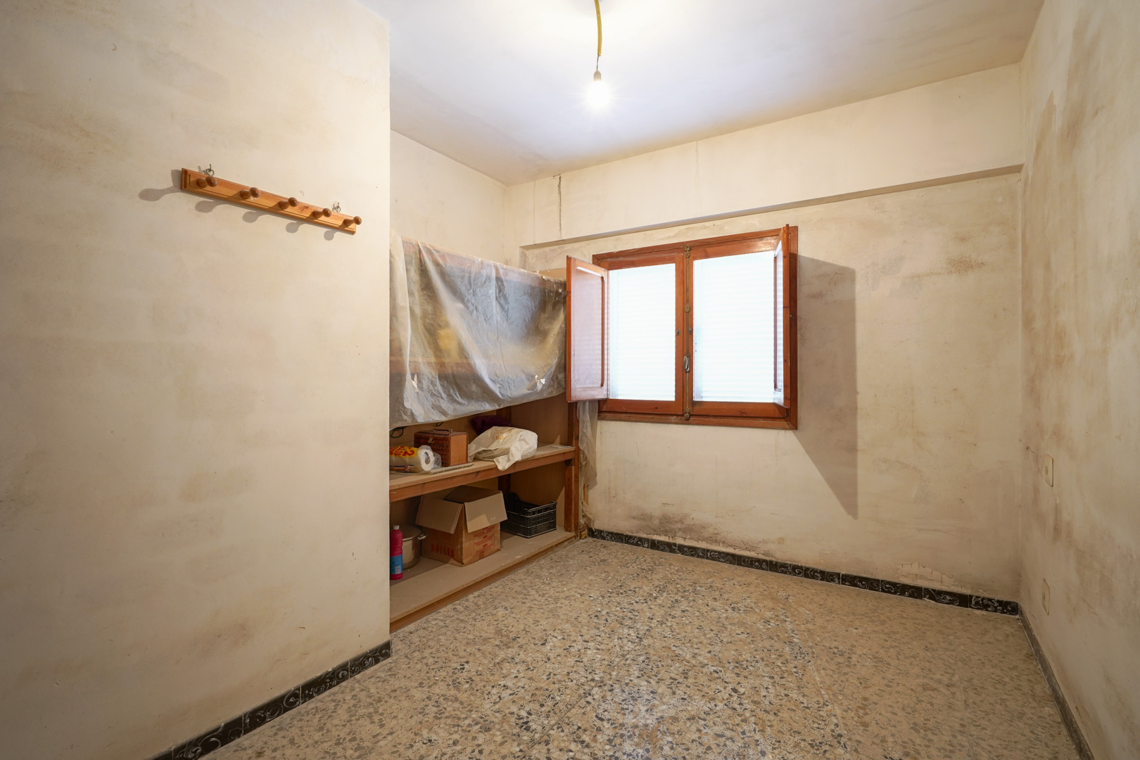 Habitación con suelos de terrazo, ventana de madera de doble hoja y estanterías integradas que requieren una reforma integral.