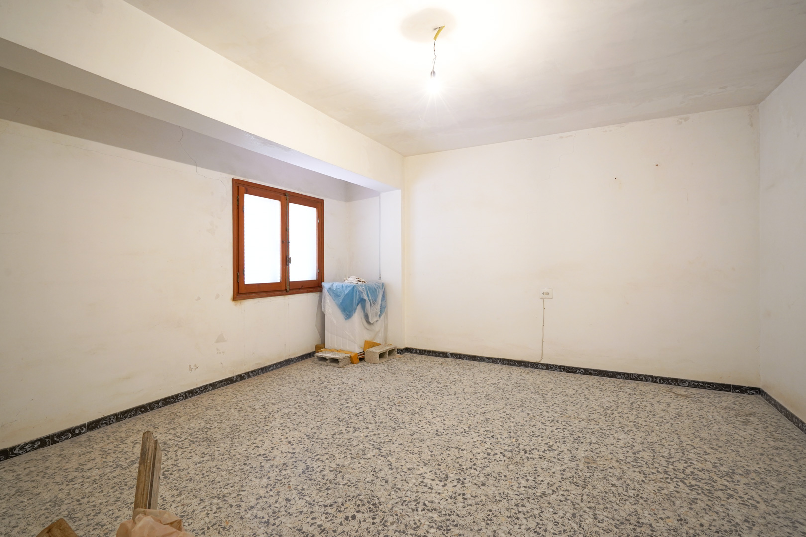 Habitación diáfana con suelos de terrazo original, paredes blancas para reformar y ventana de madera con luz natural.