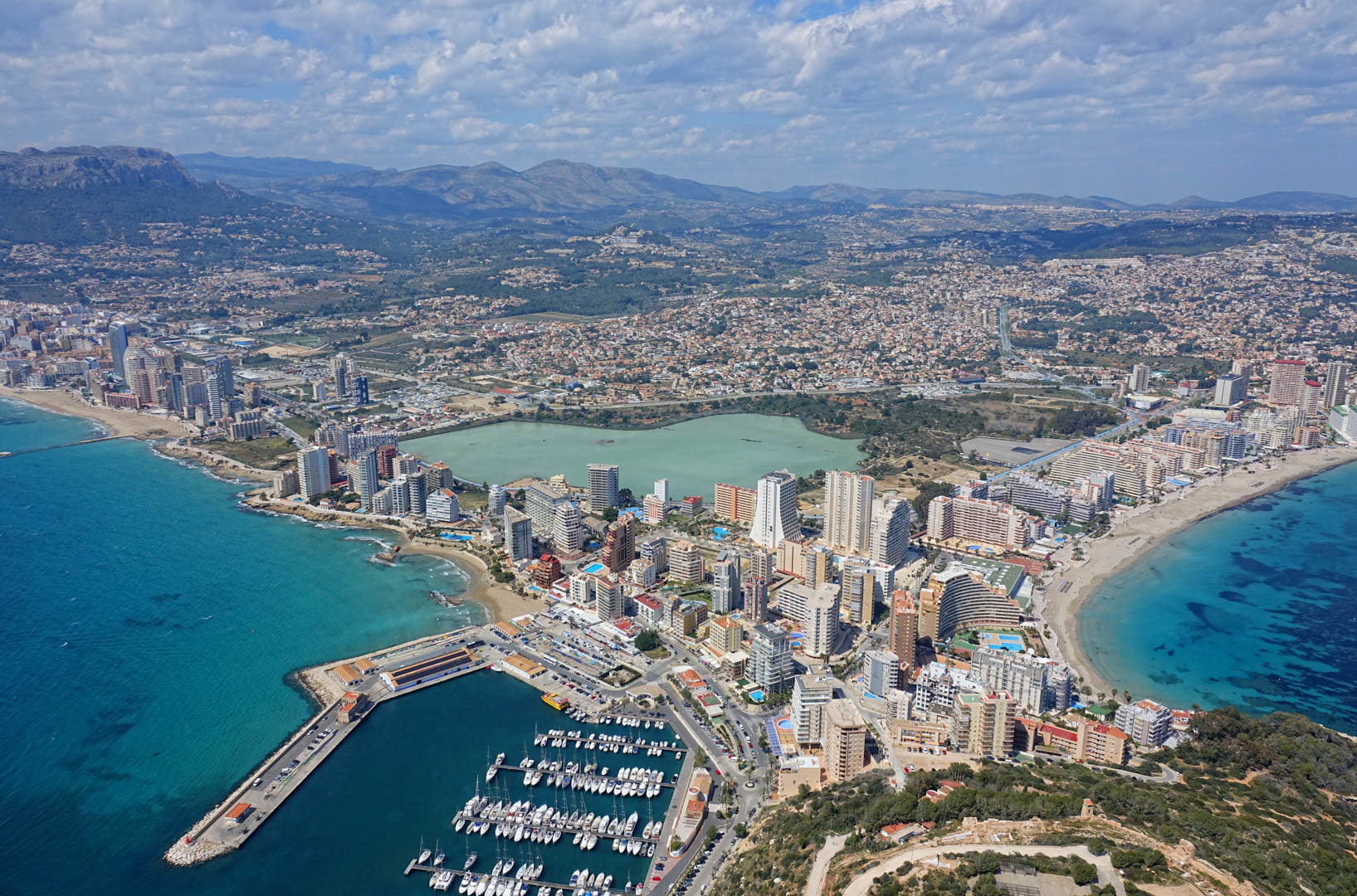 Vista aérea panorámica de Calpe que muestra el puerto deportivo, las playas de arena, la laguna de Las Salinas y las montañas de la Sierra de Bernia al fondo.