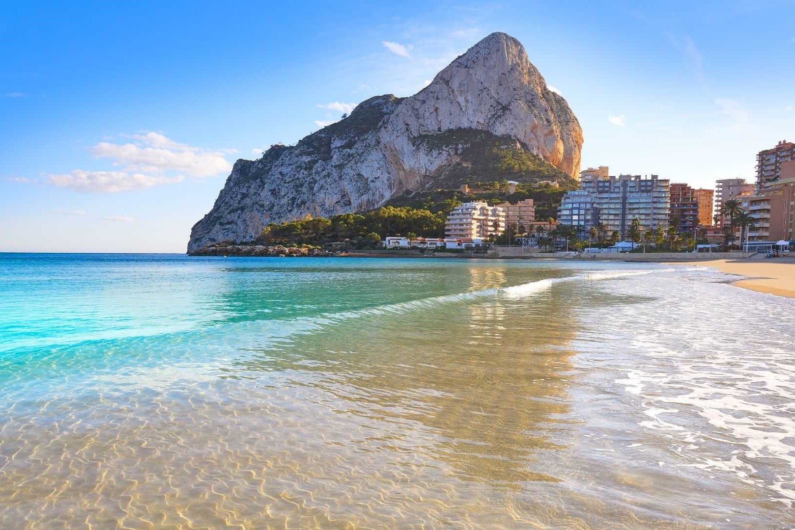 Vista panorámica del Peñón de Ifach desde la orilla de la playa en Calpe, con aguas cristalinas y edificios residenciales en primera línea de costa.