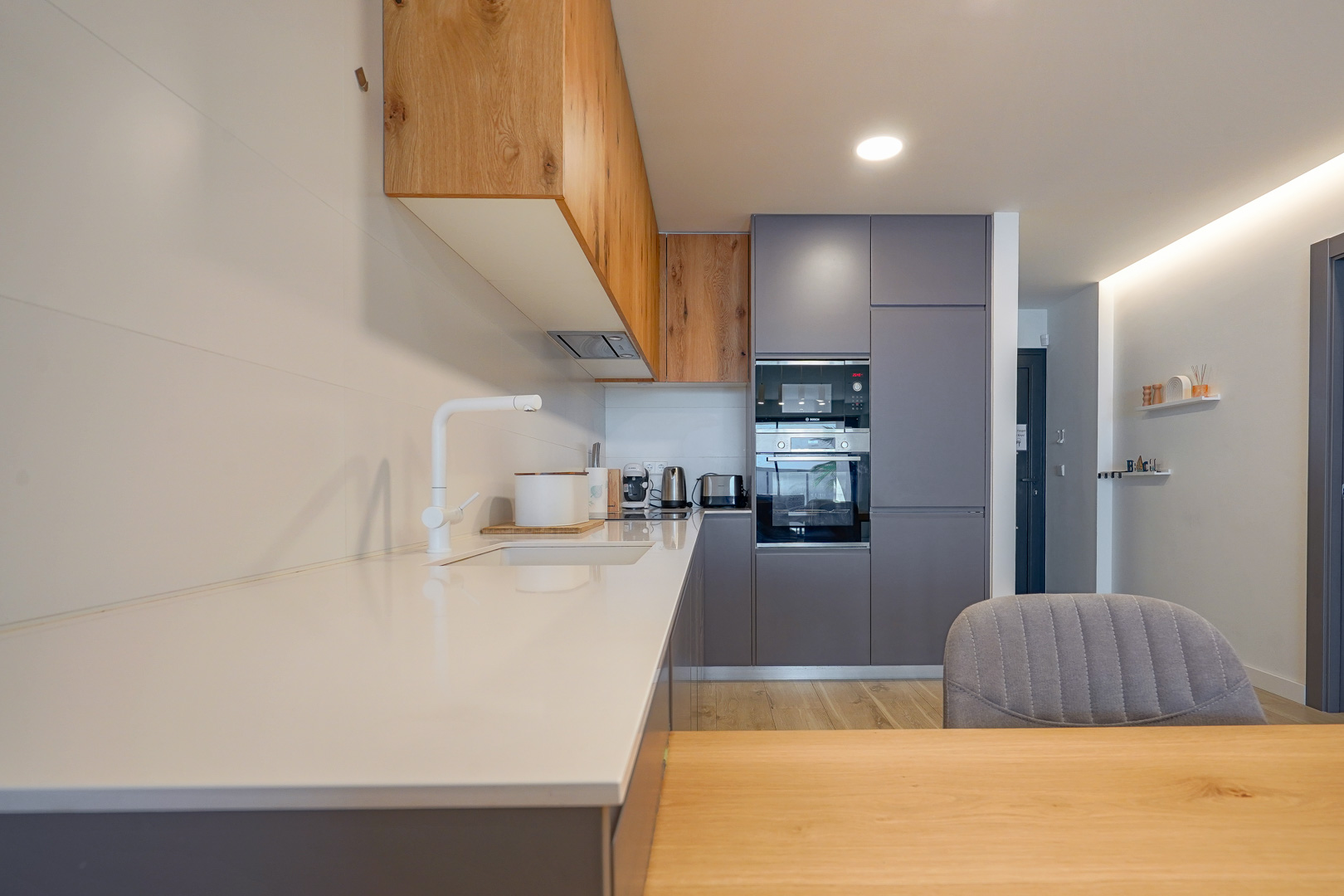 Cocina moderna con encimera blanca, armarios en gris y madera natural, equipada con electrodomésticos integrados y grifería blanca minimalista.