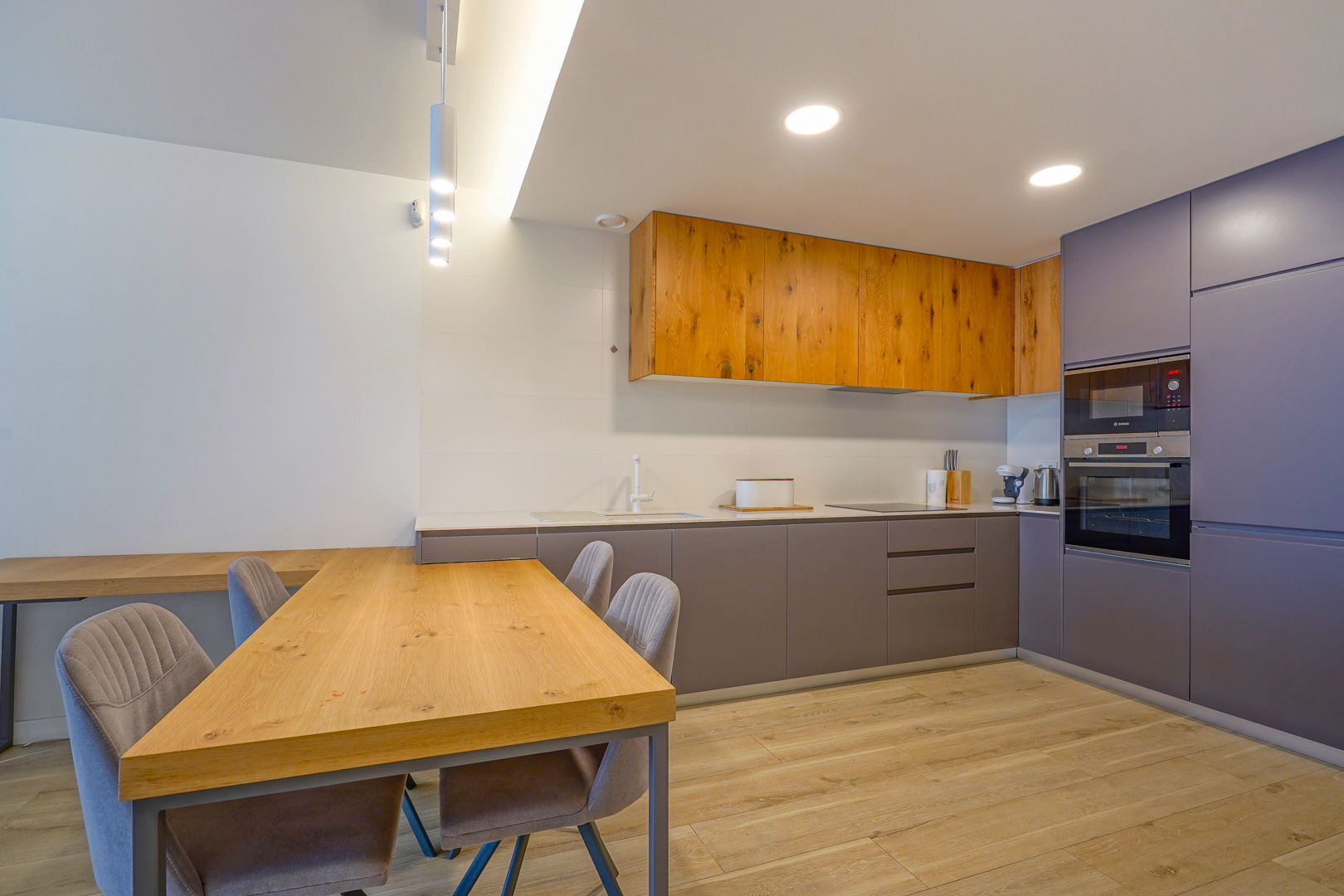 Cocina moderna de planta abierta con armarios de madera natural y gris, electrodomésticos integrados y una mesa de comedor de madera a juego con sillas tapizadas.