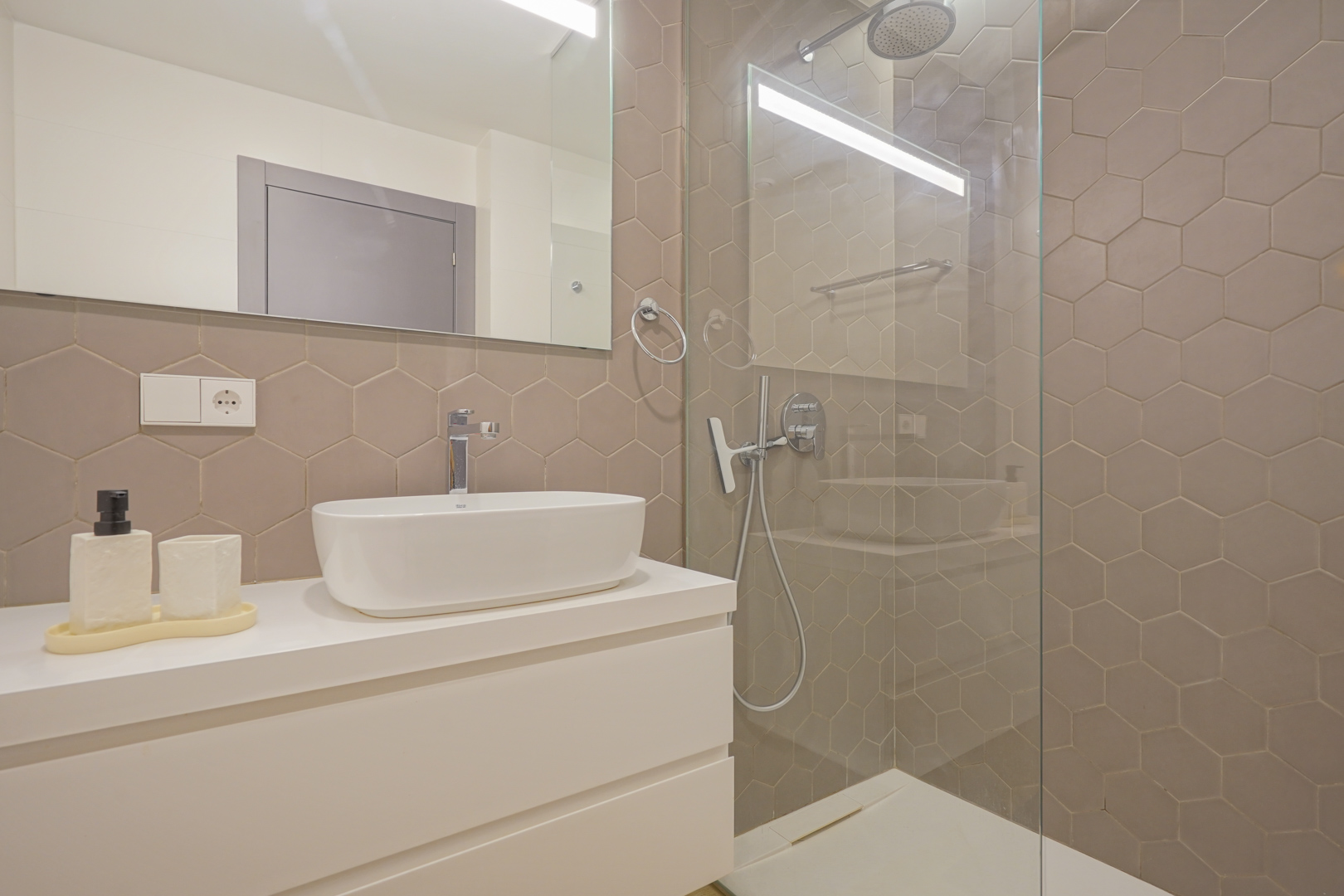 Cuarto de baño moderno con azulejos hexagonales en tono topo, lavabo sobre encimera blanca y ducha de obra con mampara de cristal fija.