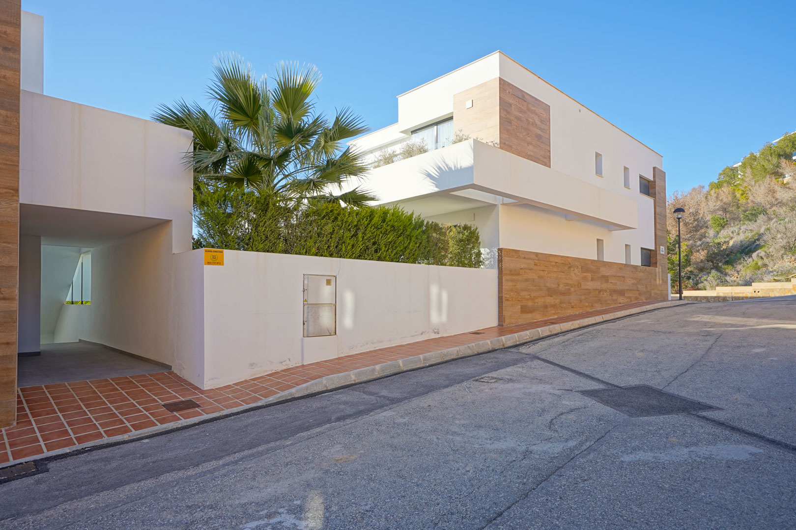 Fachada moderna del edificio en Monte Cala Gardens con acabados en blanco y gres efecto madera, incluyendo el acceso peatonal y vegetación mediterránea.
