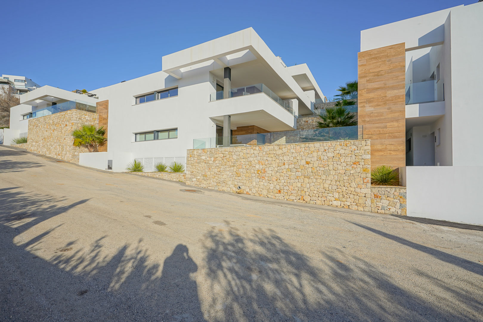 Fachada moderna en Monte Cala Gardens con muros de piedra natural, amplias terrazas con barandillas de cristal y acabados en madera bajo un cielo despejado.