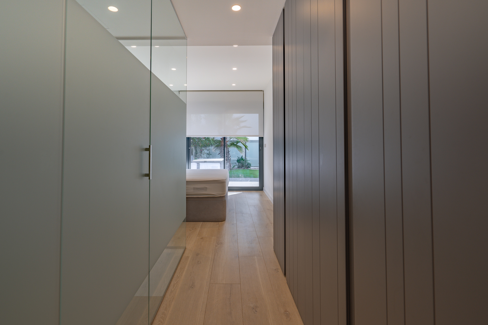 Pasillo del dormitorio principal con armarios empotrados grises y acceso al baño tras una mampara de cristal, con vistas al jardín exterior.