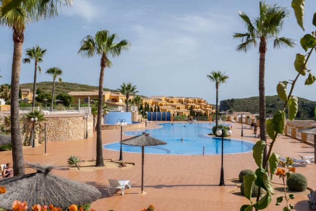 Piscina comunitaria de diseño orgánico rodeada de una amplia terraza de terracota, palmeras y sombrillas de brezo en la urbanización Monte Cala Gardens.