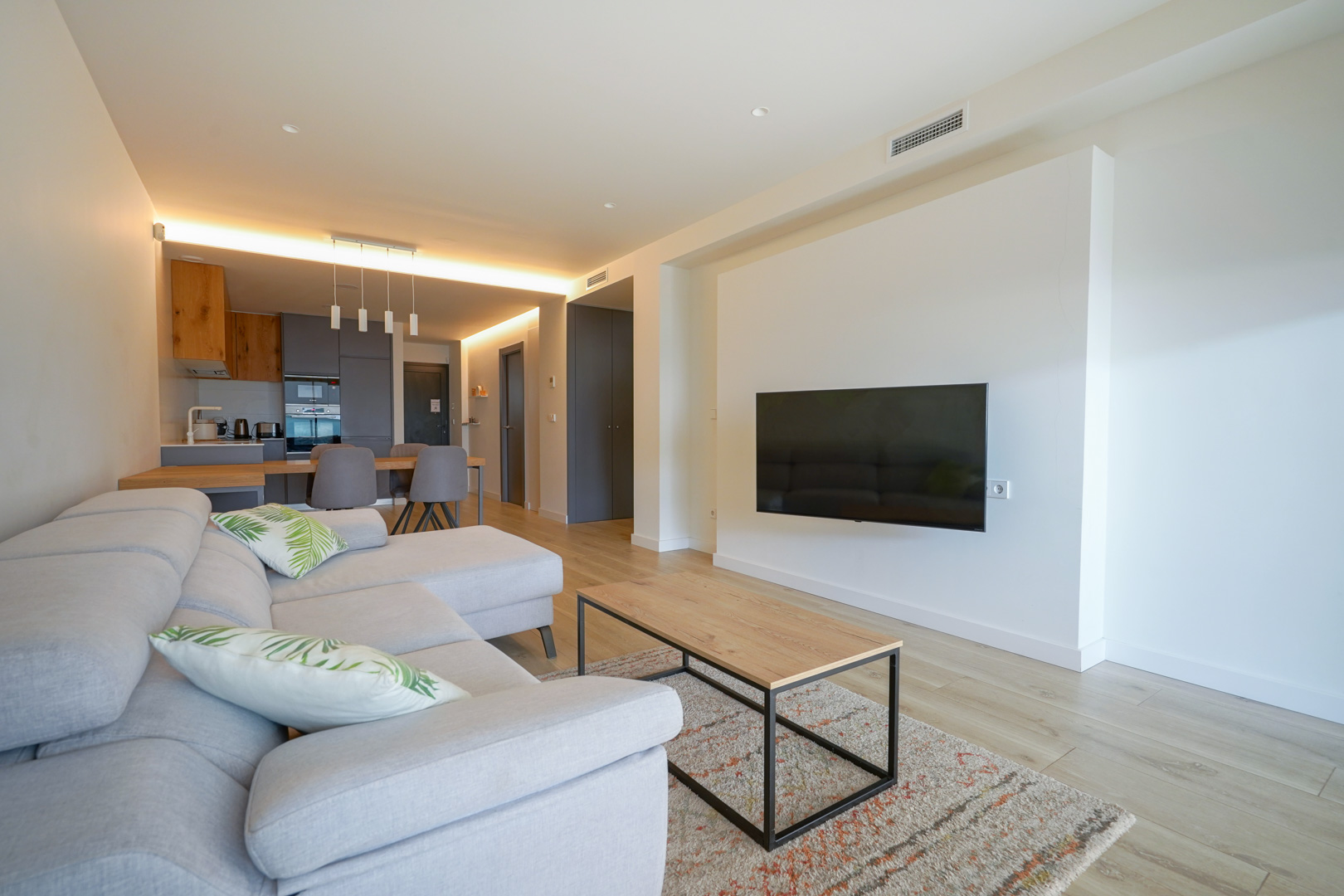 Salón de concepto abierto con televisión de pared, suelos de madera clara y cocina moderna integrada con detalles en madera e iluminación indirecta.