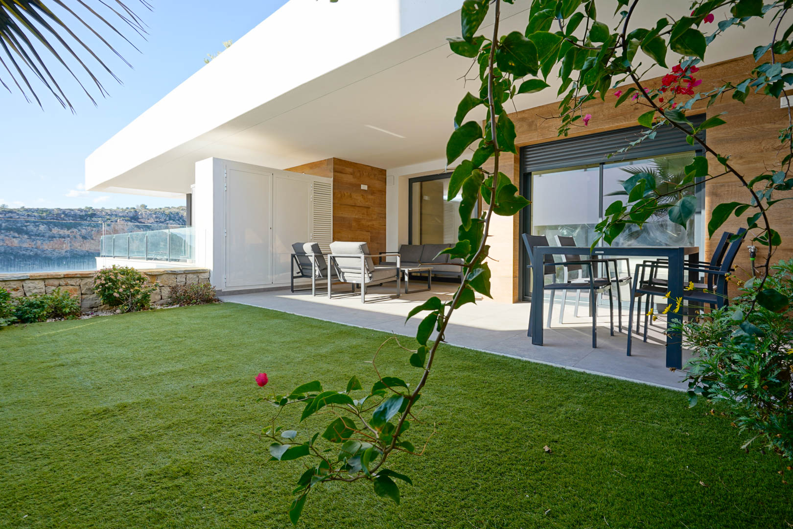 Terraza cubierta con mobiliario de exterior y jardín privado de césped artificial, delimitado por un muro de piedra natural y vistas al parque natural.