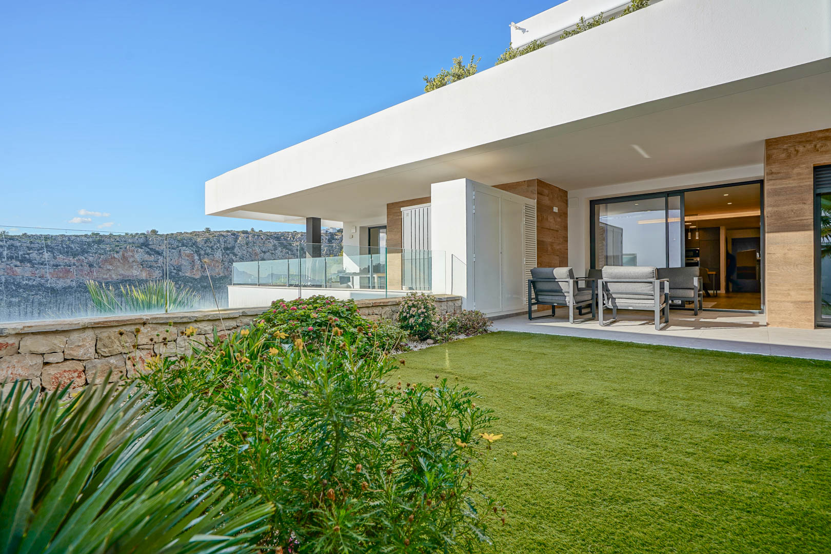 Terraza cubierta con mobiliario moderno y jardín privado de césped, delimitado por un muro de piedra natural con vistas al parque natural.
