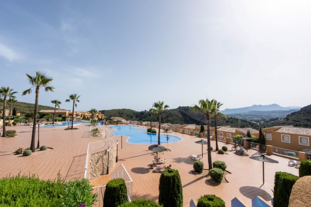 Zona comunitaria de Monte Cala Gardens con dos amplias piscinas, terraza soleada con palmeras y vistas despejadas a las montañas de Cumbre del Sol.