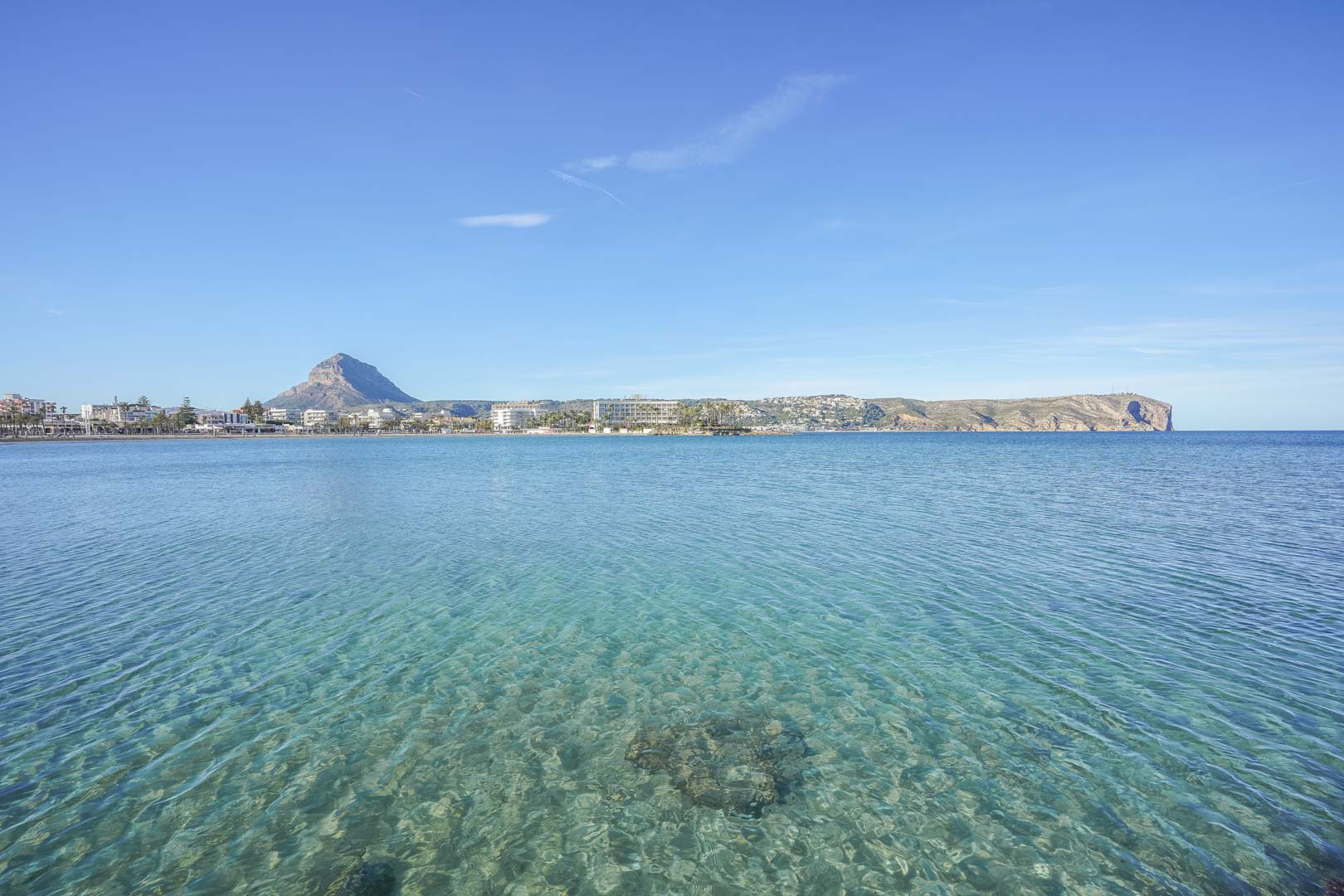 Vista panorámica de la bahía de Jávea con aguas cristalinas, mostrando el emblemático Montgó y el Cabo de San Antonio bajo un cielo despejado.