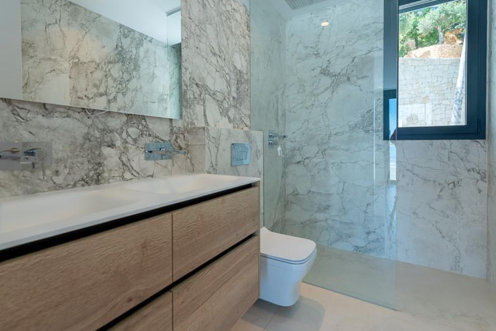 Baño moderno con revestimiento de mármol veteado, mueble de doble lavabo en madera clara y ducha a ras de suelo con mampara de cristal fija.