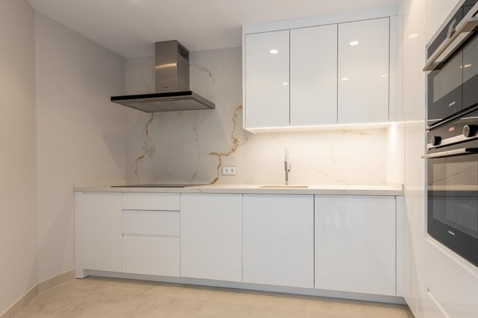 Cocina de diseño minimalista con armarios blancos sin tiradores, encimera de piedra veteada y electrodomésticos integrados de alta gama con campana de acero inoxidable.