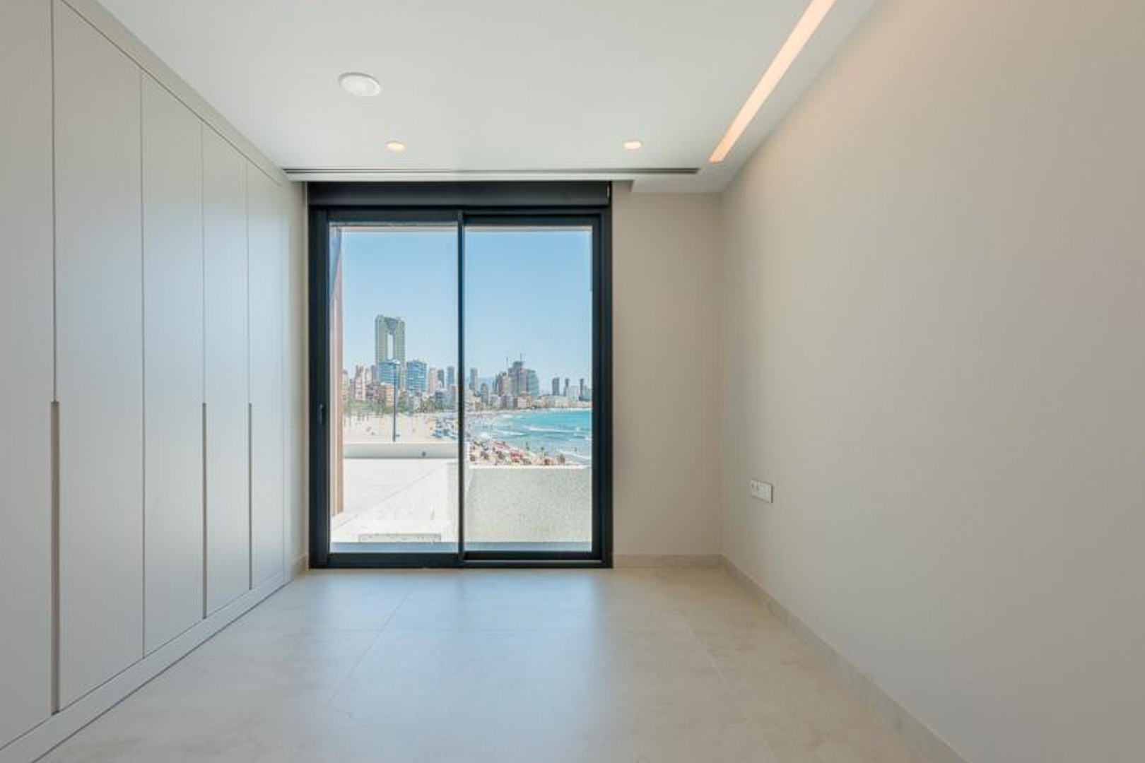 Dormitorio con armarios empotrados de suelo a techo, iluminación LED integrada y vistas directas a la playa de Poniente y el skyline de Benidorm.