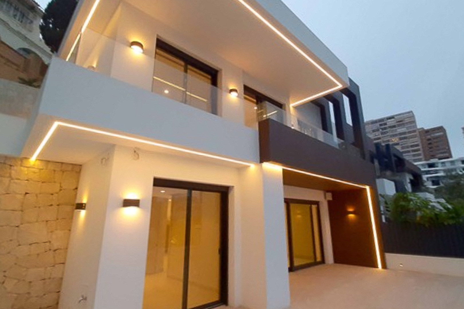 Fachada exterior de diseño contemporáneo con iluminación LED perimetral, barandillas de cristal y grandes ventanales que conectan con el interior.