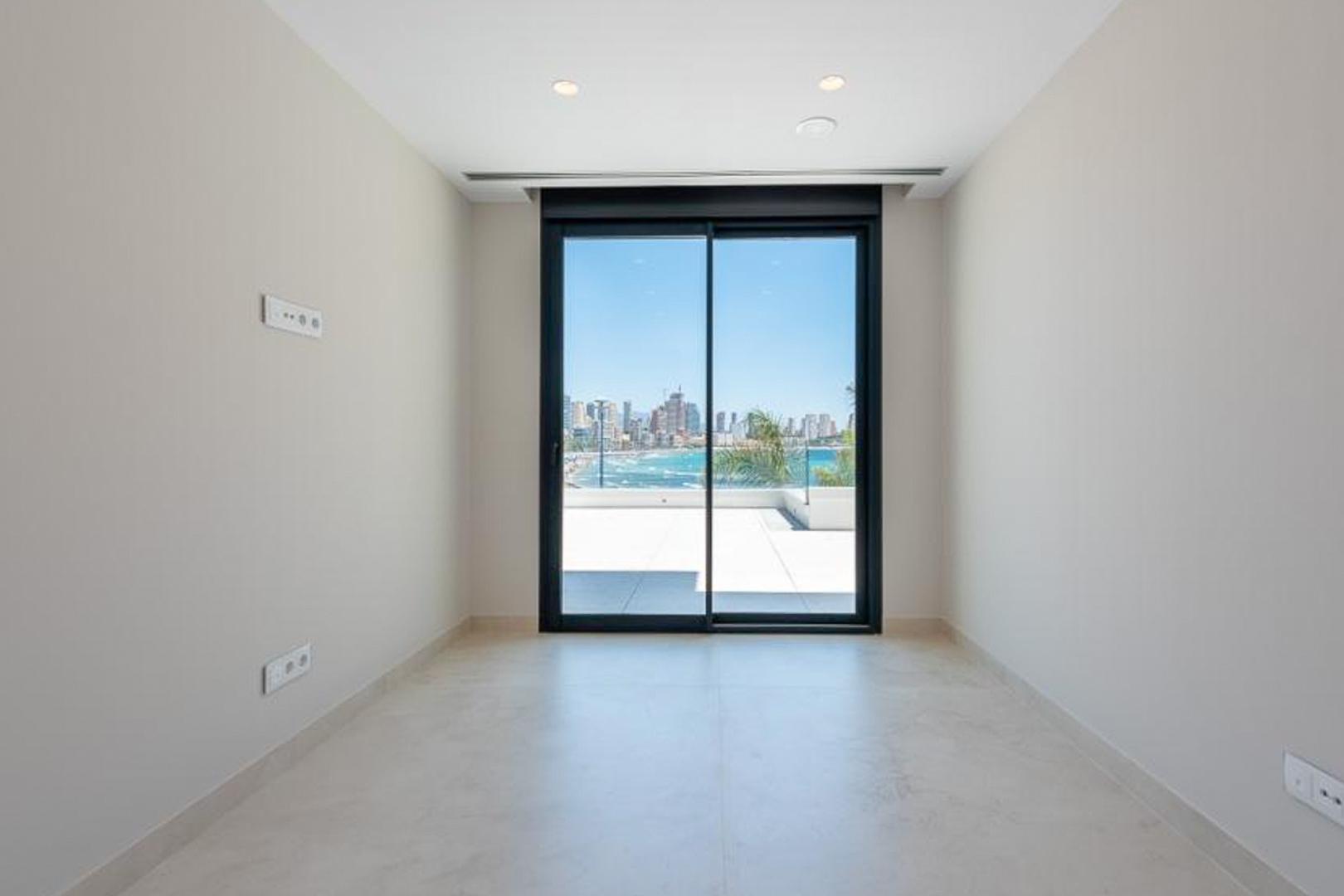Habitación luminosa con suelos porcelánicos, iluminación empotrada y acceso directo a la terraza mediante un gran ventanal con vistas al mar y al skyline de Benidorm.
