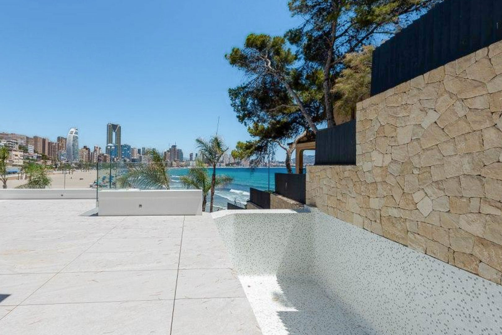 Terraza exterior con suelo de piedra clara y piscina de gresite blanco, ofreciendo vistas panorámicas a la playa y al skyline de Benidorm tras un muro de mampostería natural.