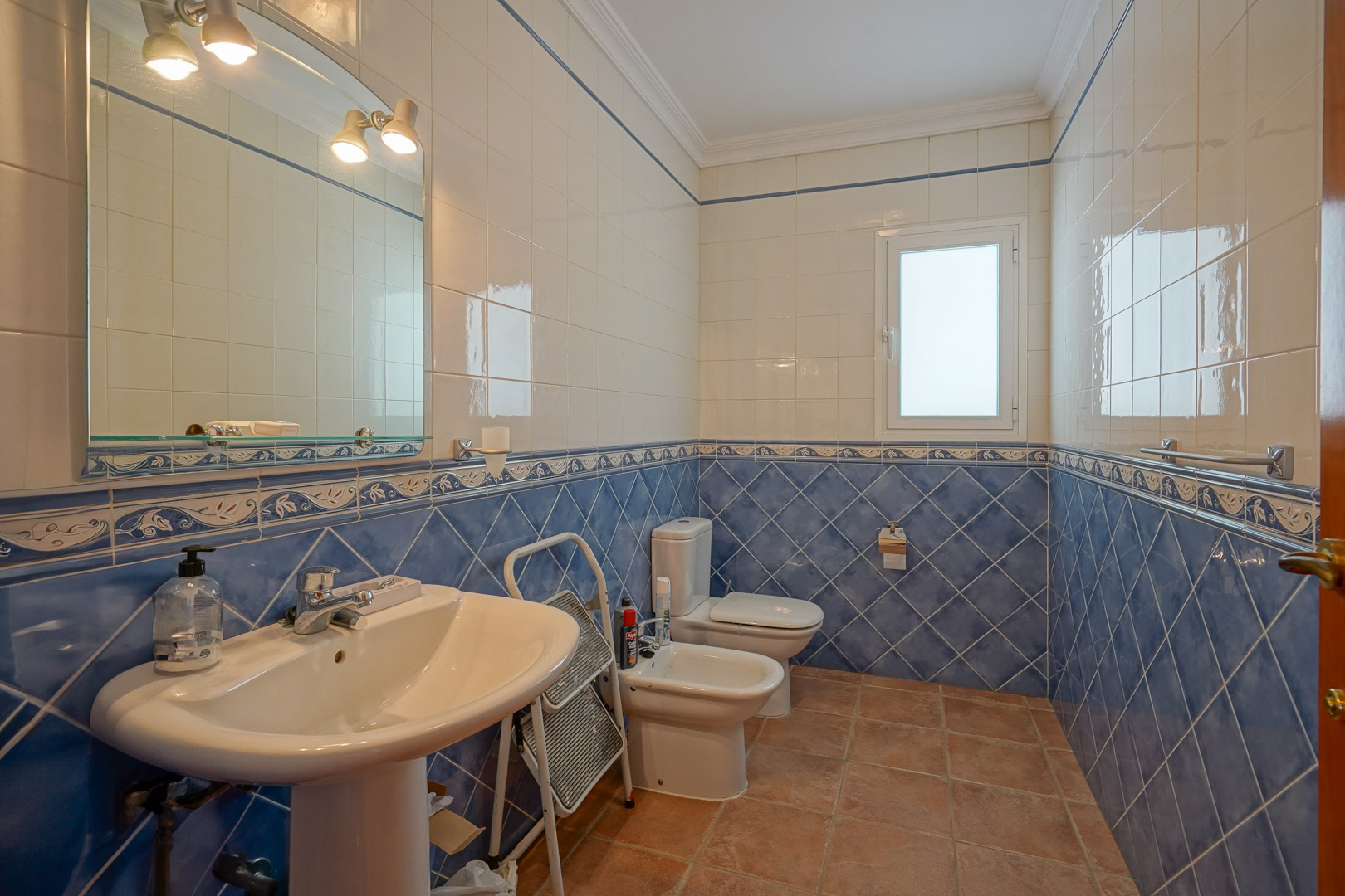 Baño con azulejos bicolores en azul y blanco, suelos de terracota y sanitarios clásicos que incluyen lavabo de pie, bidé e inodoro bajo una iluminación integrada.