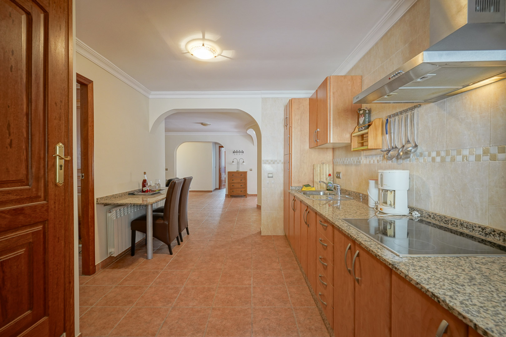Cocina funcional con mobiliario de madera, encimeras de granito y placa vitrocerámica. Incluye una barra de desayuno integrada y suelos de gres que conectan con la vivienda mediante arcos.