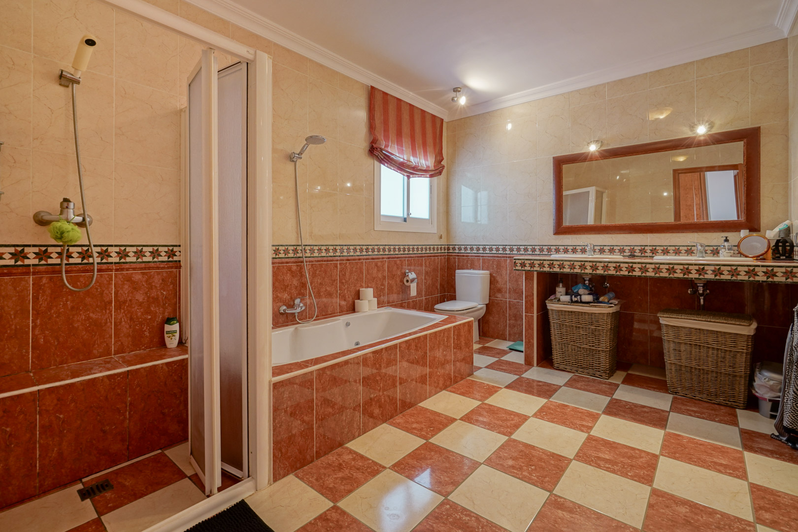 Cuarto de baño con azulejos en tonos crema y rojizos, equipado con bañera encastrada, ducha independiente y encimera de doble seno con espejo de marco de madera.