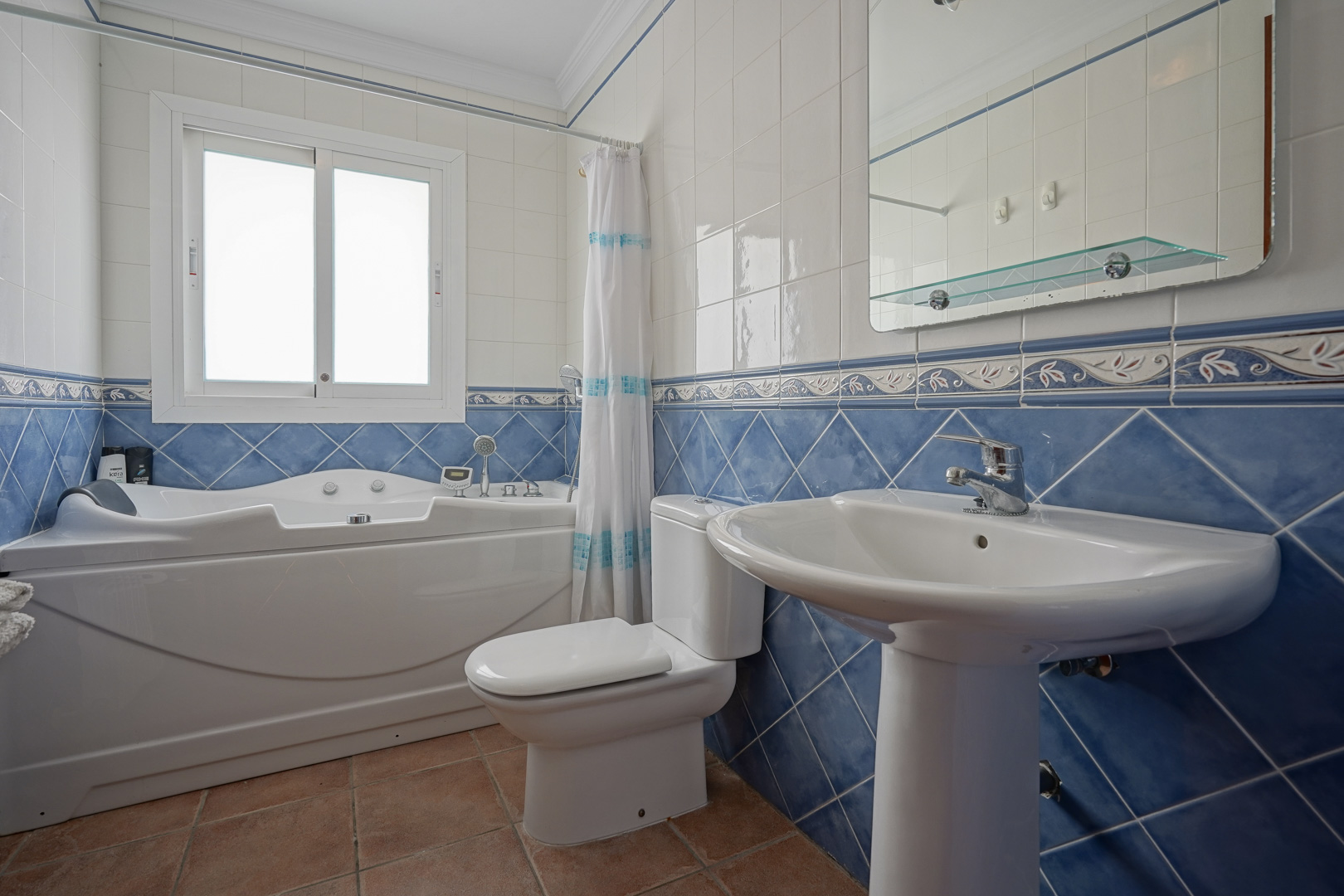 Cuarto de baño con bañera de hidromasaje, lavabo de pedestal y azulejos en tonos azules y blancos. Incluye suelo de terracota y ventana para luz natural.