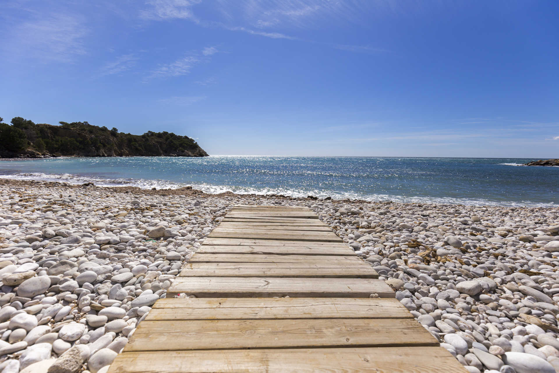Playa de guijarros blancos en Altea con una pasarela de madera que se extiende hacia el mar Mediterráneo, rodeada de un entorno natural costero.
