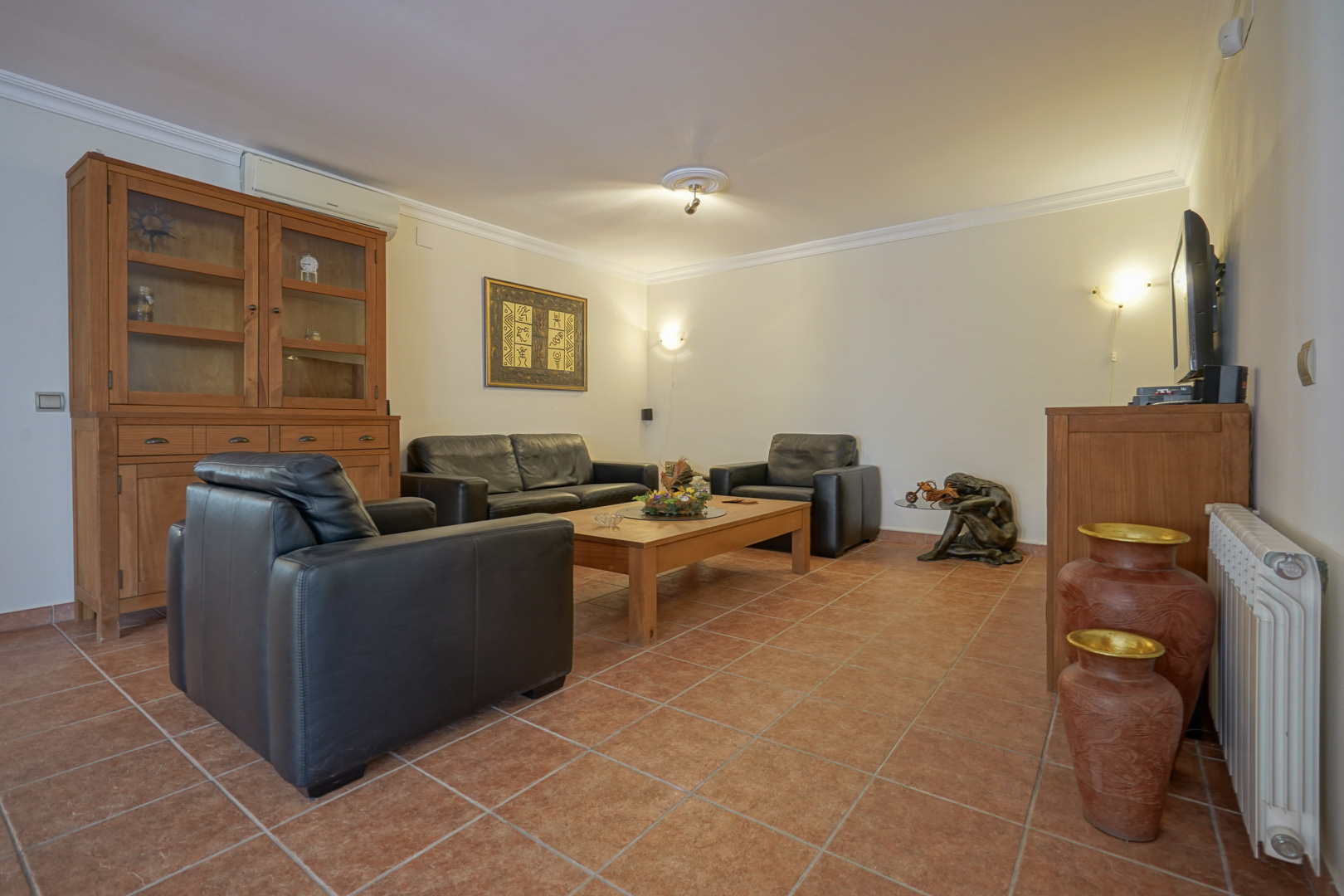 Salón con sofás de cuero negro, muebles de madera maciza y suelos de gres rústico. Incluye aire acondicionado, radiador y una vitrina clásica de madera.