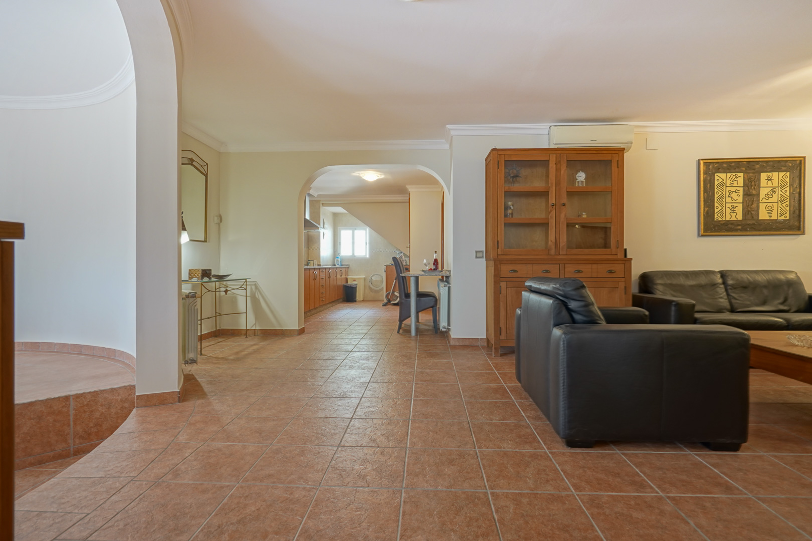 Salón con sofás de cuero negro y suelos de gres cerámico, conectado mediante un arco a una cocina equipada con mobiliario de madera y zona de comedor.