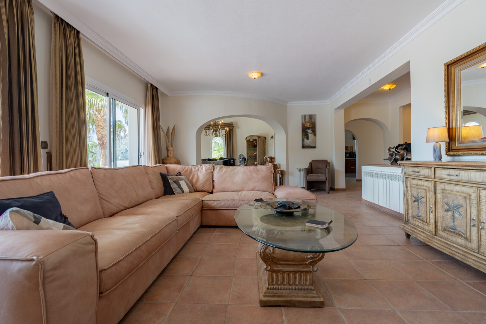Salón con suelos de terracota, un gran sofá rinconero y mesa de centro de cristal. Los arcos conectan con el comedor y el pasillo bajo techos con molduras elegantes.