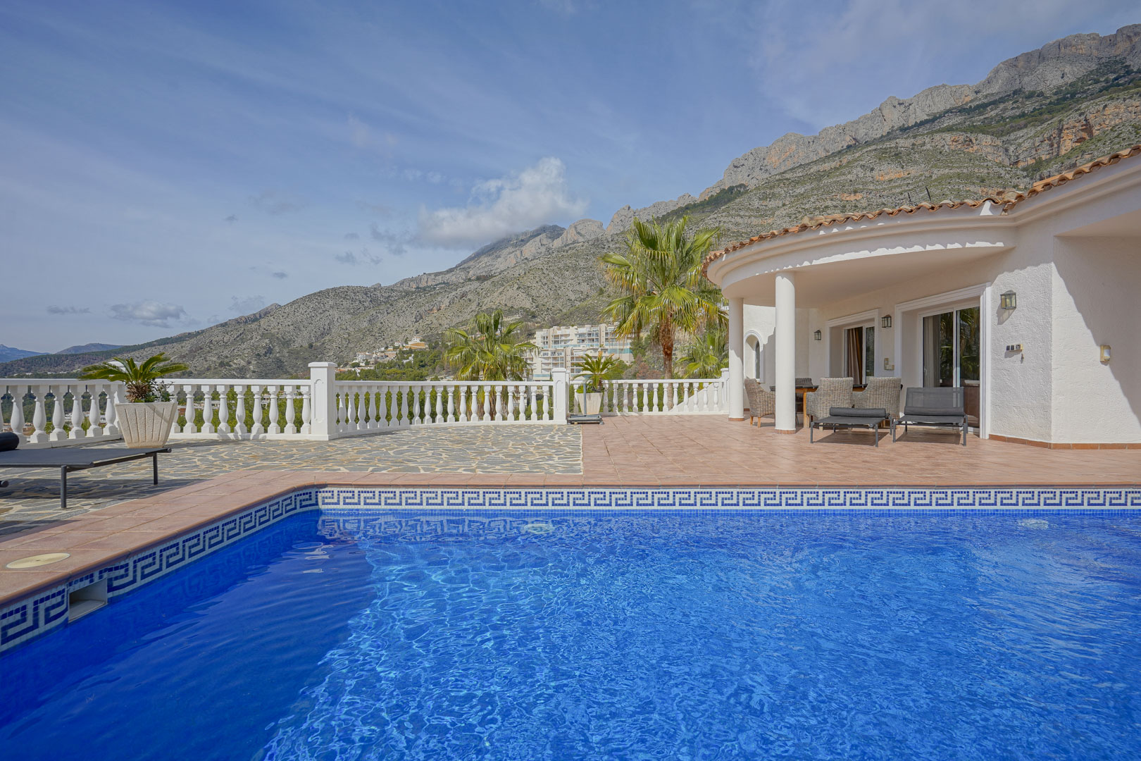 Terraza con piscina de mosaico azul y porche cubierto, ofreciendo vistas despejadas a la Sierra de Bernia y el paisaje montañoso de Altea.