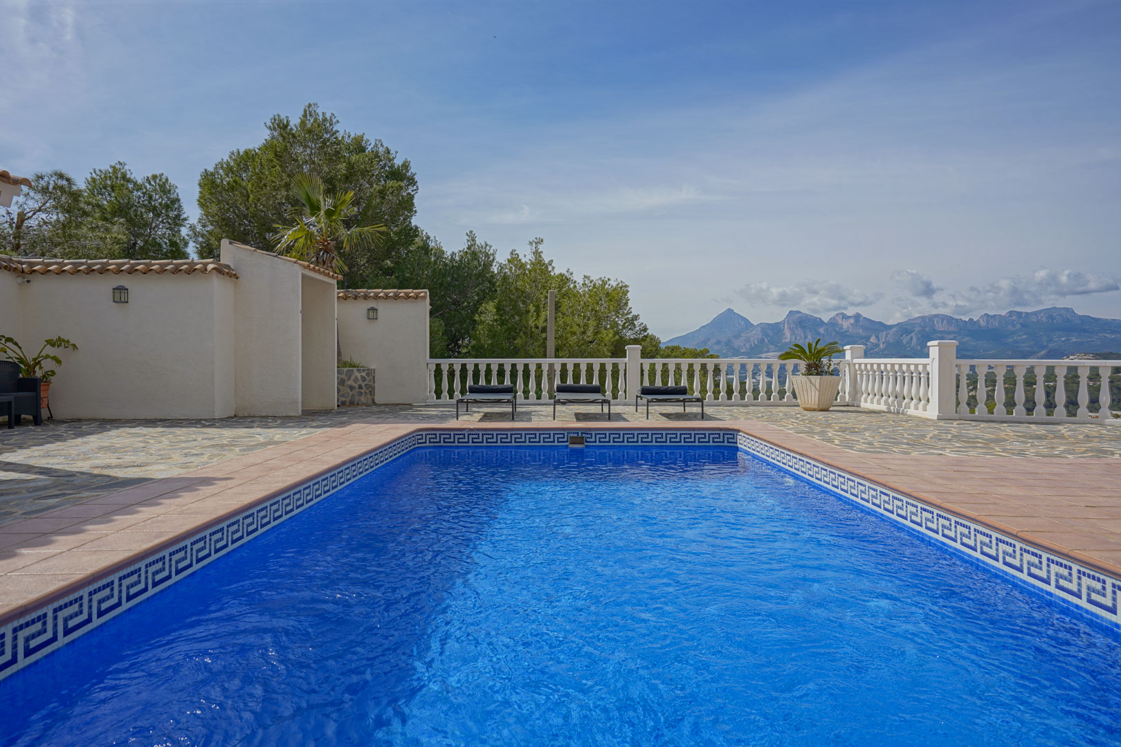 Terraza de la piscina con pavimentos de piedra natural y balaustrada blanca, ofreciendo vistas despejadas a las montañas del Puig Campana y la Sierra Aitana.