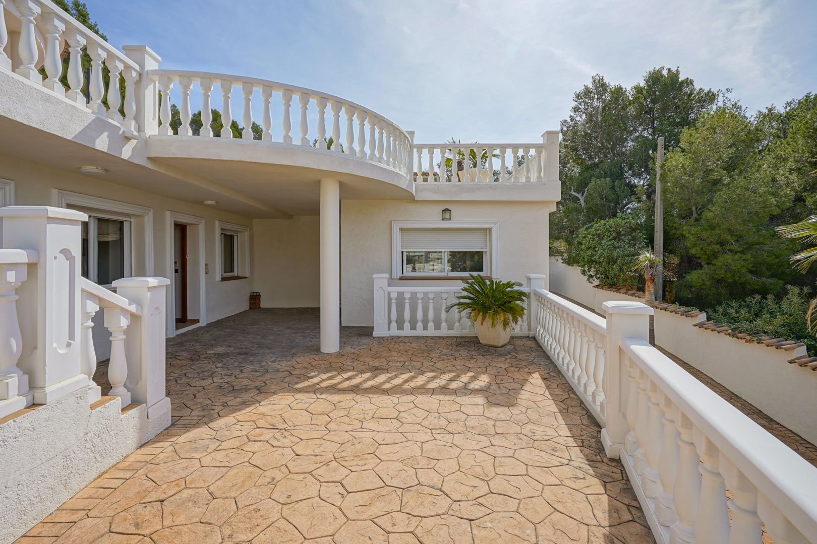 Terraza exterior con suelo de piedra estampada y balaustradas blancas clásicas, situada bajo el porche curvo que conecta los niveles de la villa.