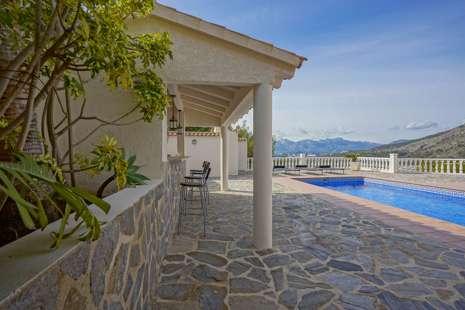 Terraza exterior con suelo de piedra natural, cocina de verano con barra y vistas panorámicas a las montañas junto a la piscina privada.