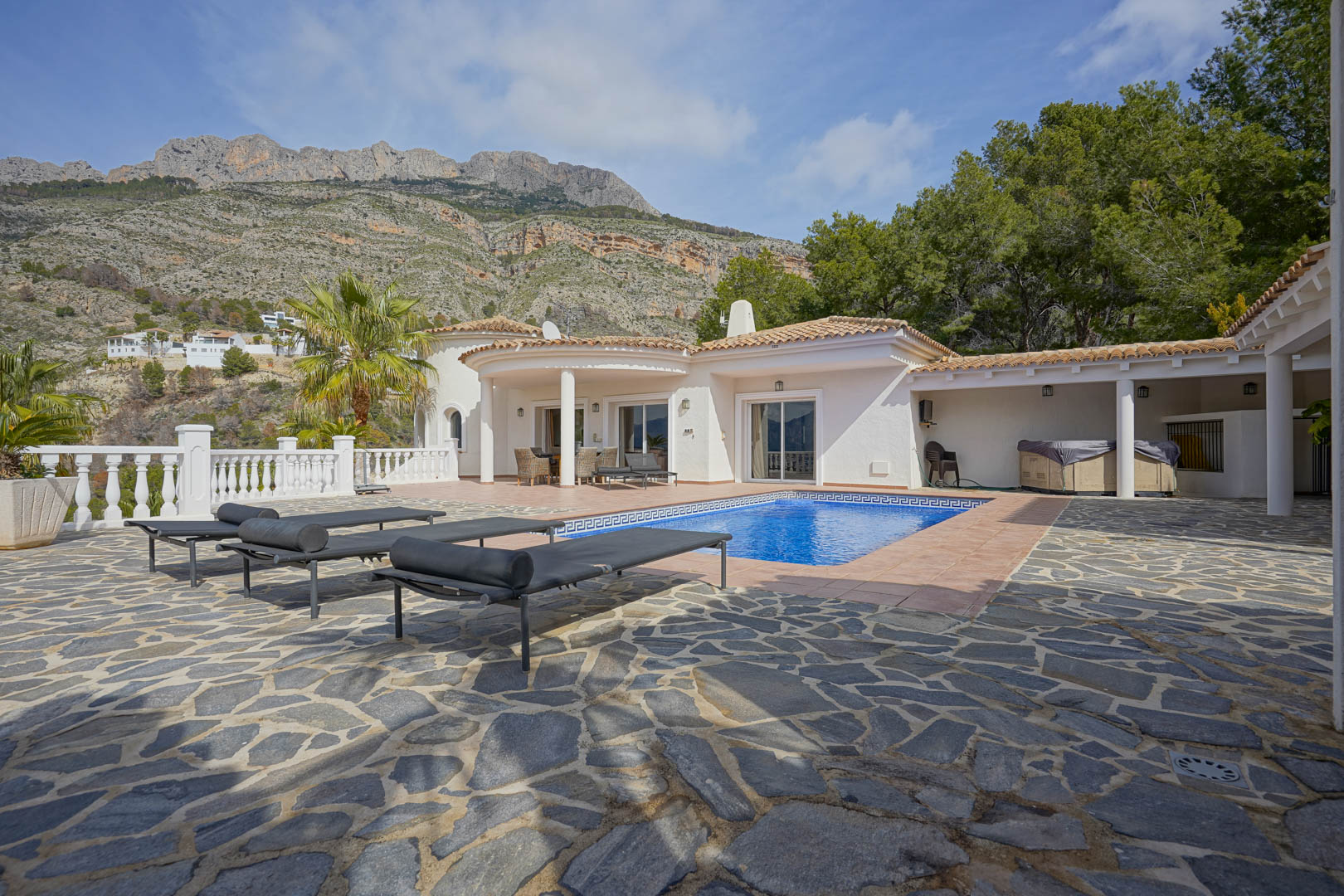 Terraza principal con piscina y pavimentación de piedra natural, destacando las vistas frontales a la Sierra de Bernia y la arquitectura blanca de estilo mediterráneo.