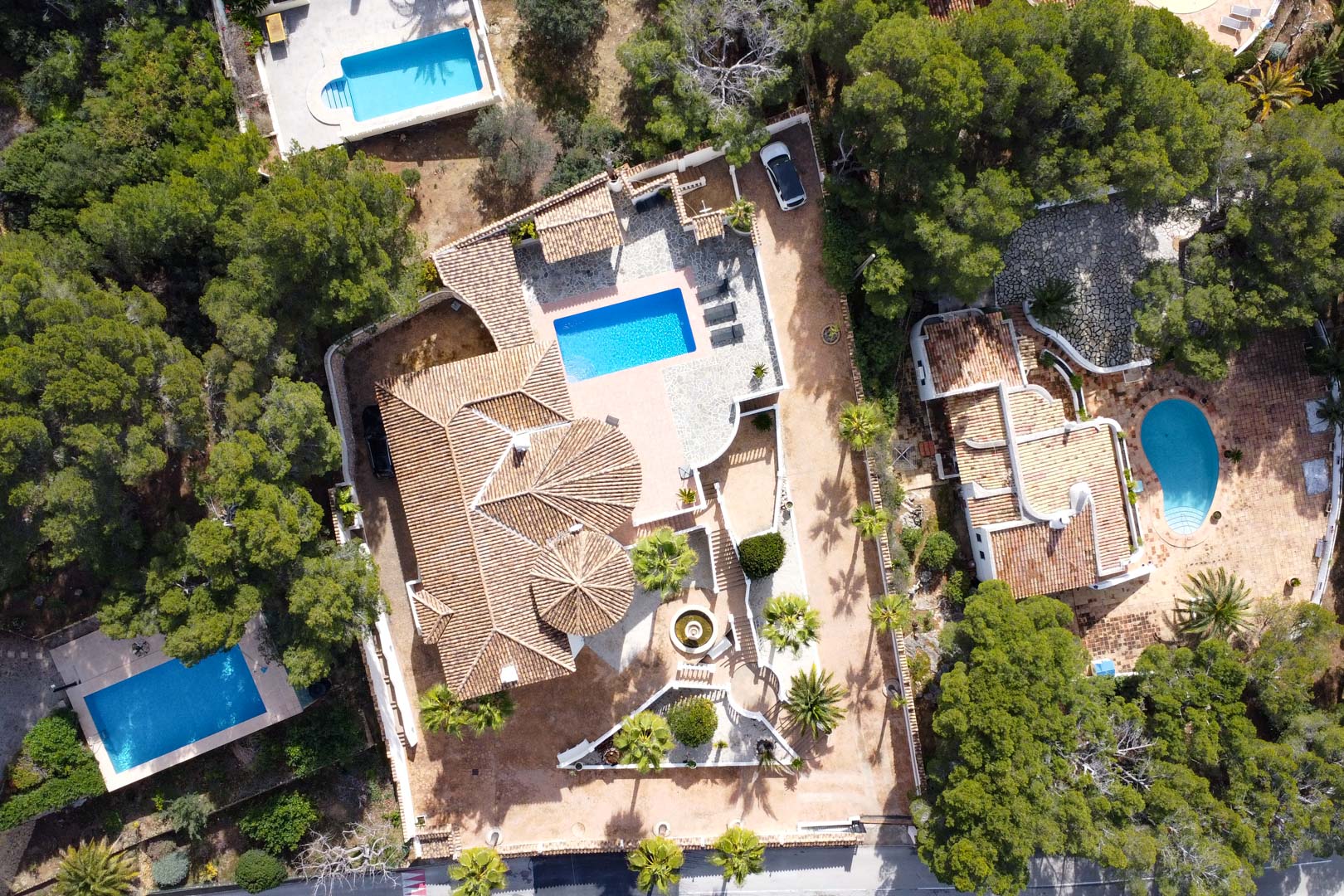 Vista aérea cenital de la villa que muestra la arquitectura de tejados de teja, la piscina rectangular y las amplias terrazas rodeadas de vegetación mediterránea.