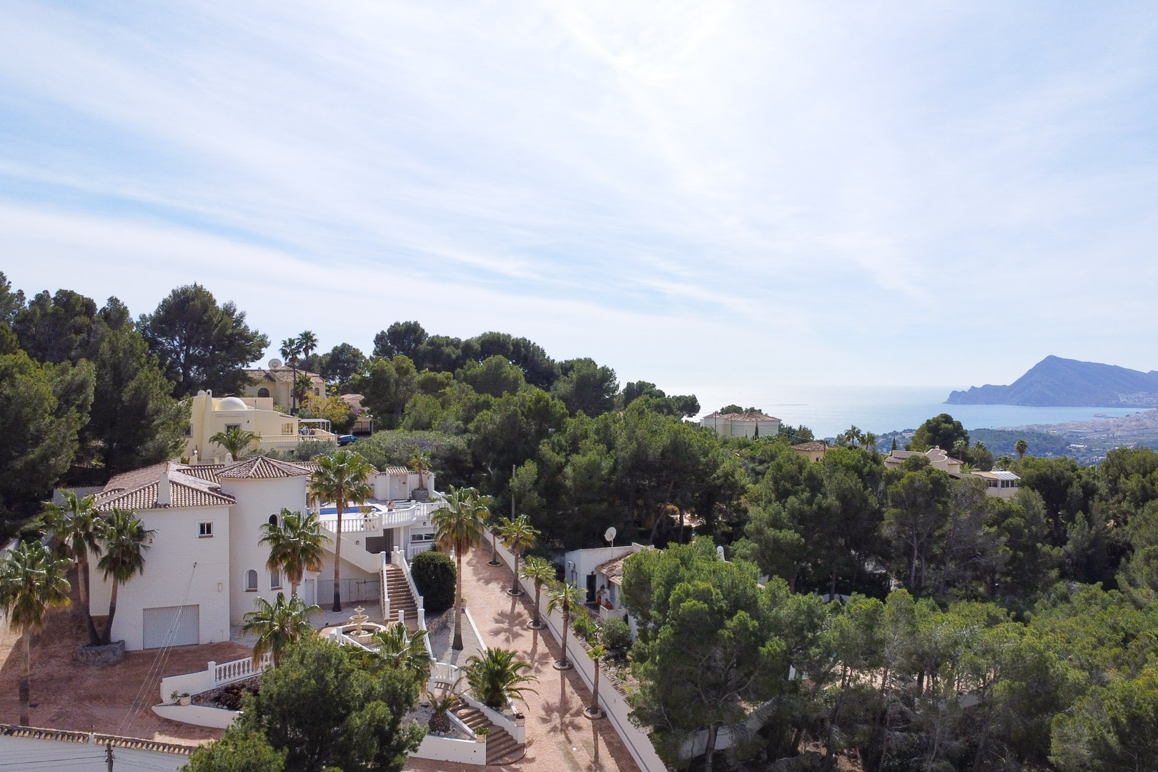 Vista aérea de la villa blanca con arquitectura mediterránea, rodeada de pinos y palmeras, ofreciendo vistas panorámicas hacia la costa de Altea y el mar.