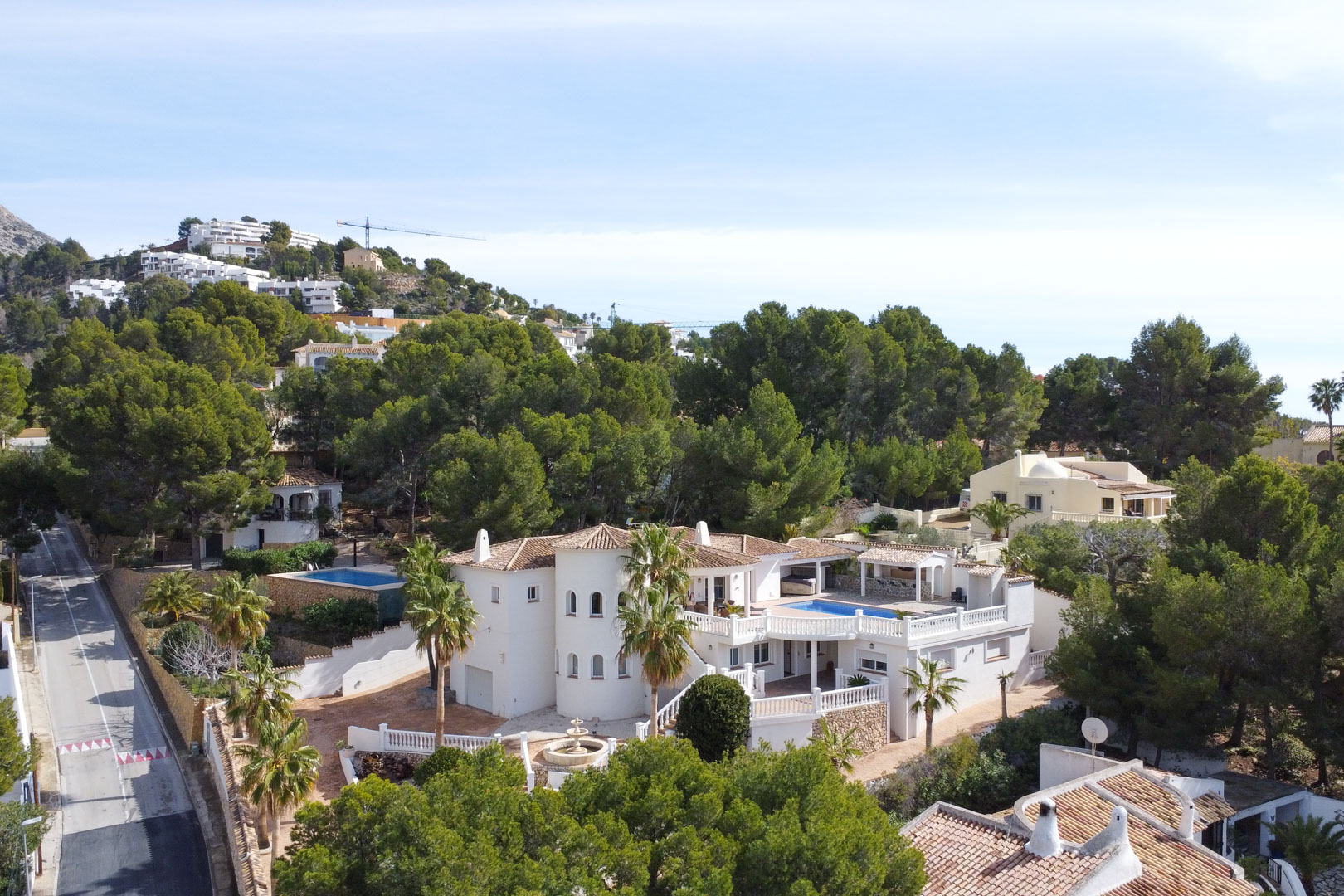 Vista aérea de la villa mediterránea con su fachada blanca, torreón circular y terraza con piscina, rodeada de un frondoso bosque de pinos en Altea.