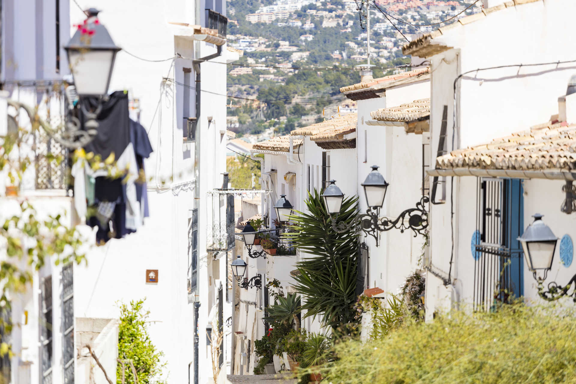 Vista de una calle estrecha en el casco antiguo de Altea, con casas blancas tradicionales, farolas de hierro forjado y vistas a la ladera montañosa.