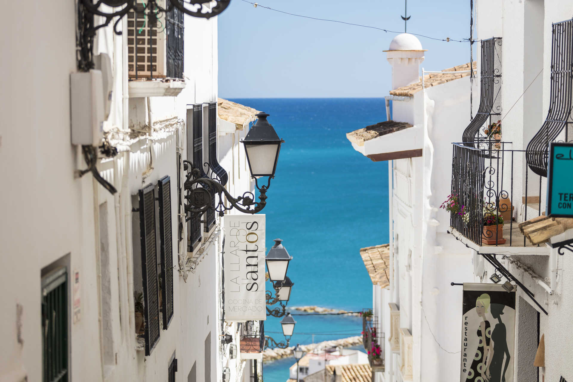 Vista de una calle estrecha en el casco antiguo de Altea, con fachadas blancas, farolas de hierro forjado y vistas panorámicas al mar Mediterráneo.
