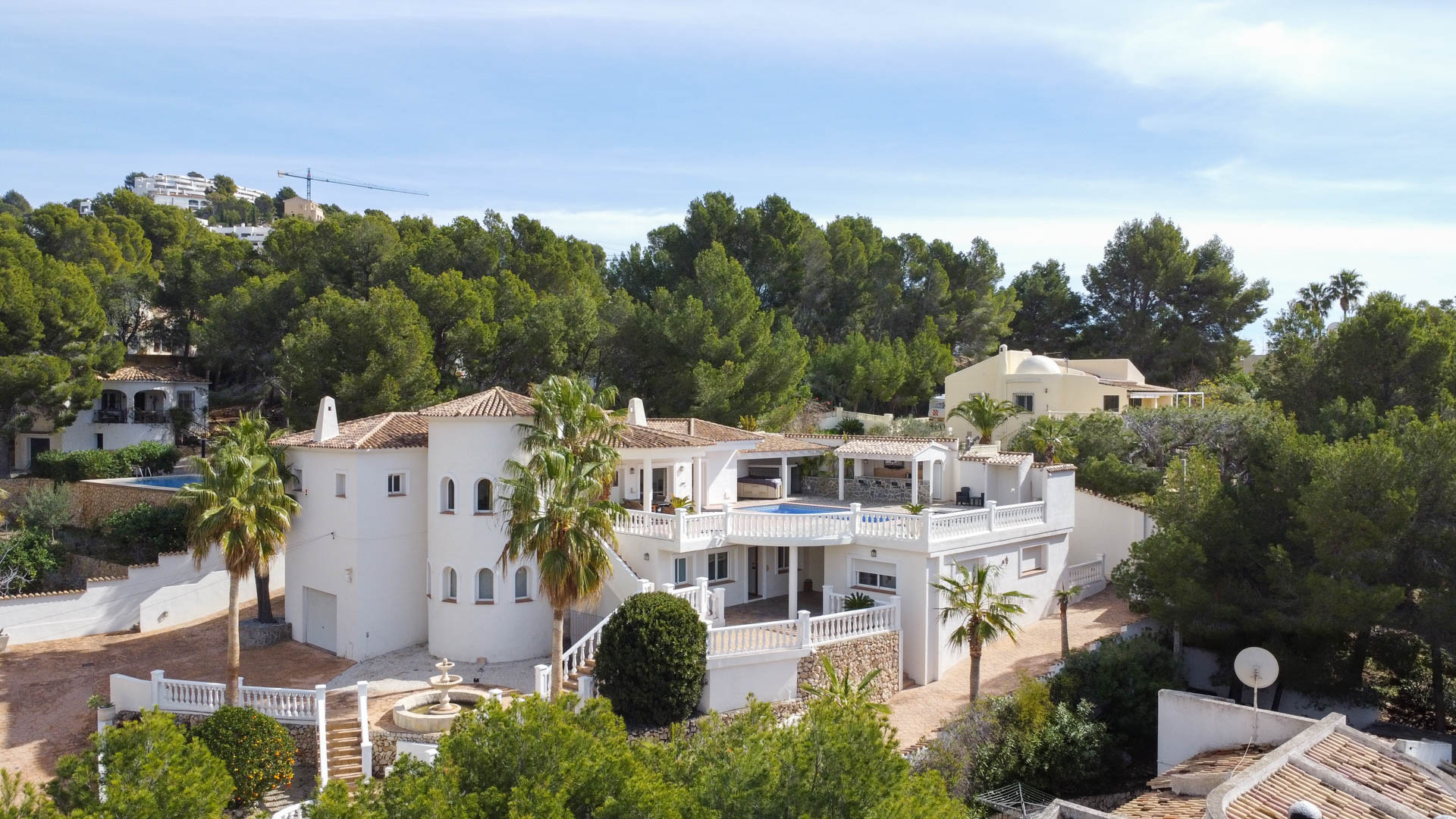 Vista exterior de la villa mediterránea con fachada blanca, torre circular y terrazas con balaustradas. Incluye piscina elevada y entorno rodeado de pinos.
