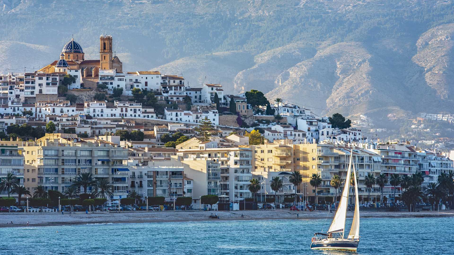 Vista panorámica del casco antiguo de Altea con su icónica cúpula azul, la Sierra de Bernia al fondo y un velero navegando frente a la costa mediterránea.