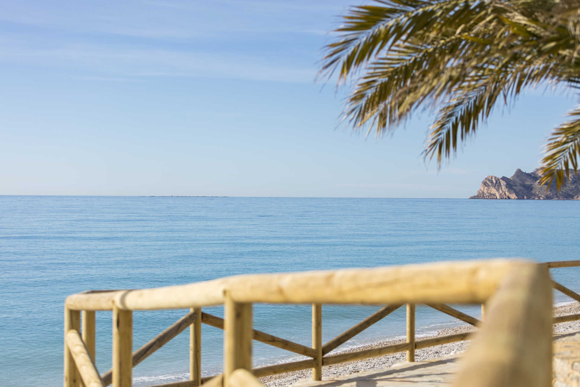 Vista panorámica del mar Mediterráneo desde el paseo marítimo de Altea, con barandillas de madera, una playa de guijarros y acantilados al fondo bajo un cielo despejado.