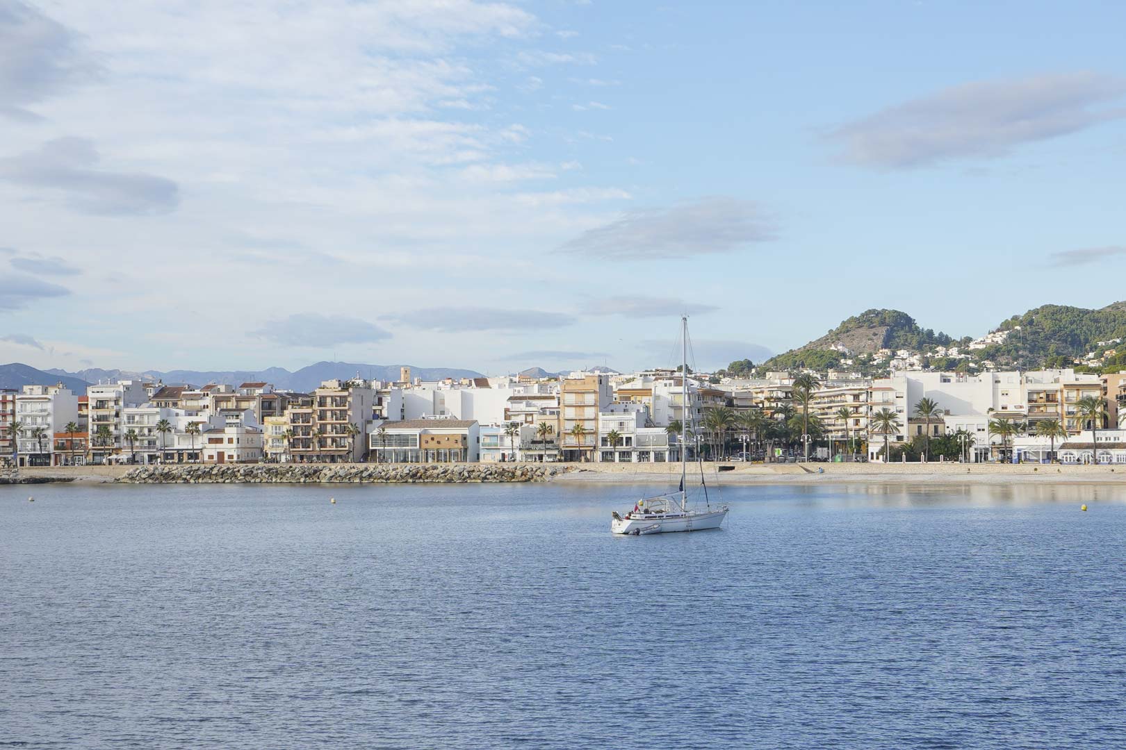 Vista panorámica de la costa de Jávea con un velero en aguas tranquilas, destacando la arquitectura blanca y las montañas al fondo.