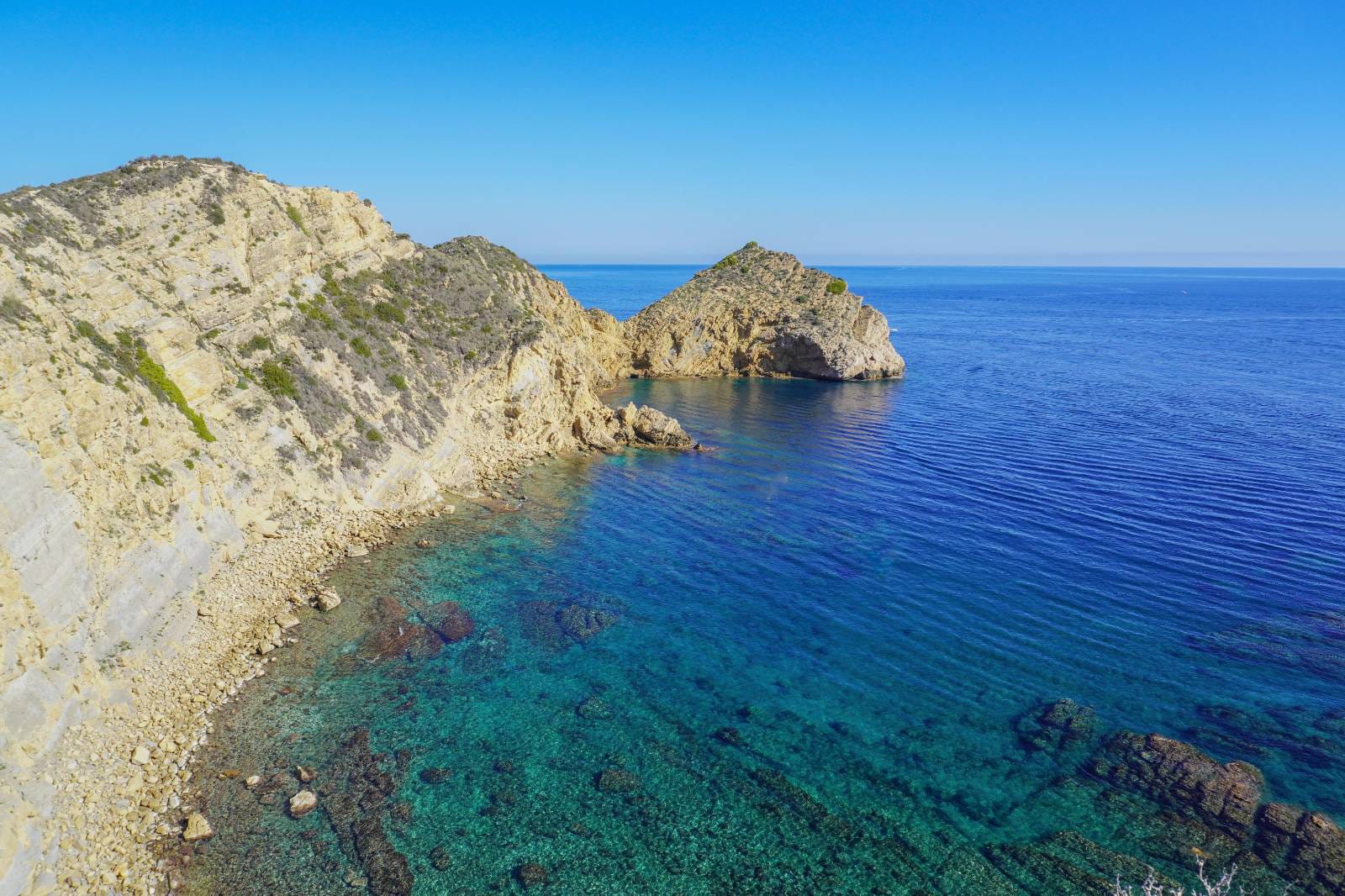 Vista panorámica de los acantilados de Jávea con aguas cristalinas de color turquesa y fondos rocosos bajo un cielo despejado.