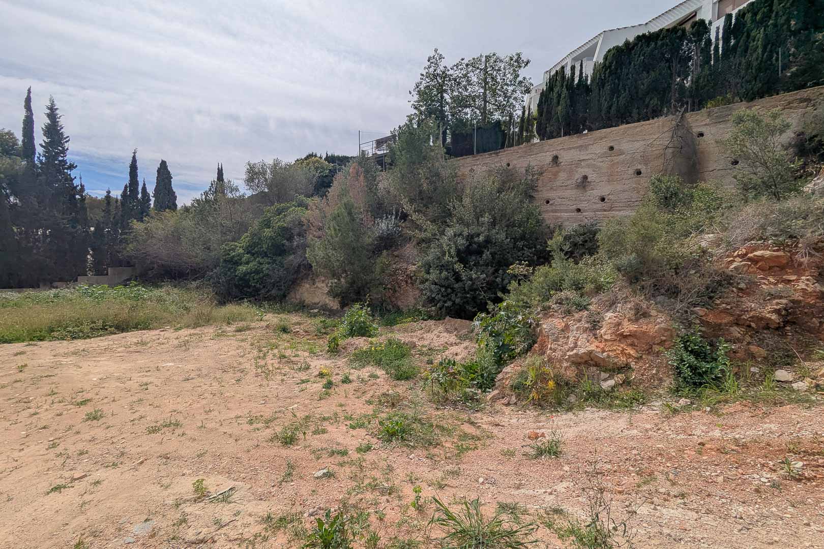 Parcela elevada en La Corona con muro de contención de hormigón y vegetación mediterránea autóctona. Terreno preparado para el inicio de un proyecto de construcción a medida.
