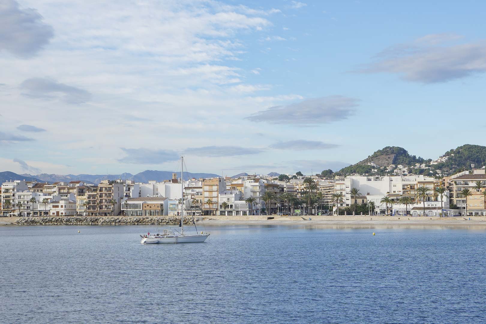 Vista panorámica del puerto de Jávea con veleros fondeados frente al paseo marítimo y las montañas de la Costa Blanca al fondo.