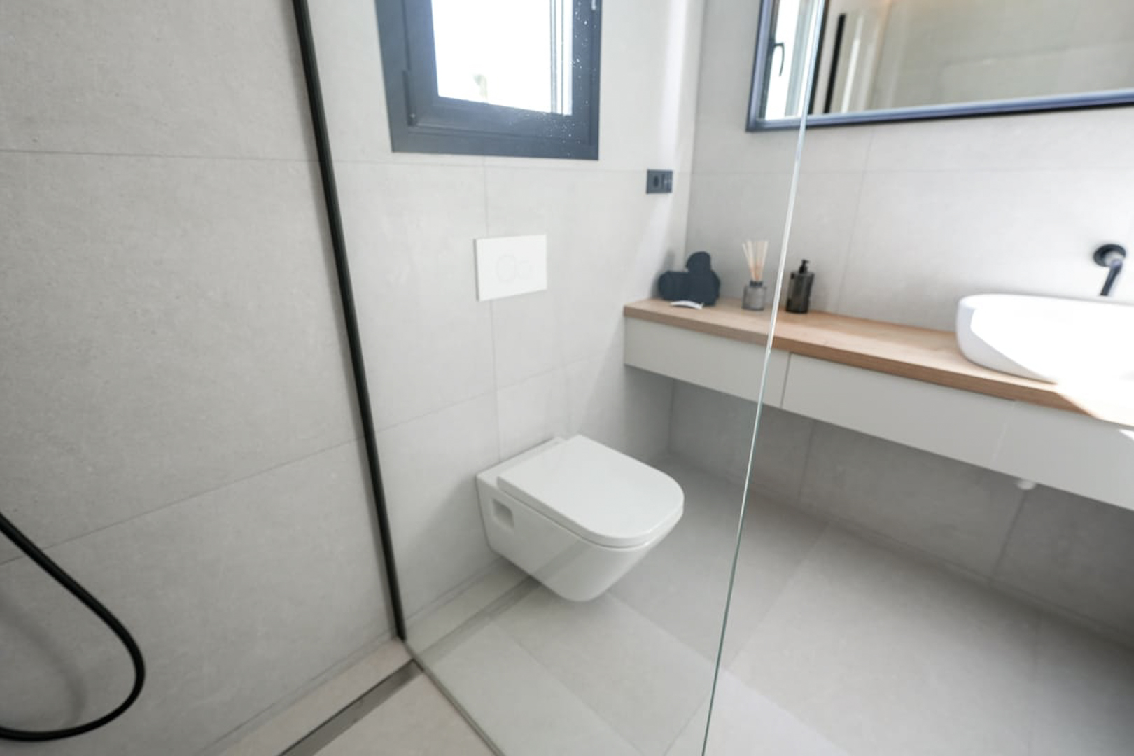 Detalle de baño moderno con azulejos grises, inodoro suspendido y encimera de madera clara.