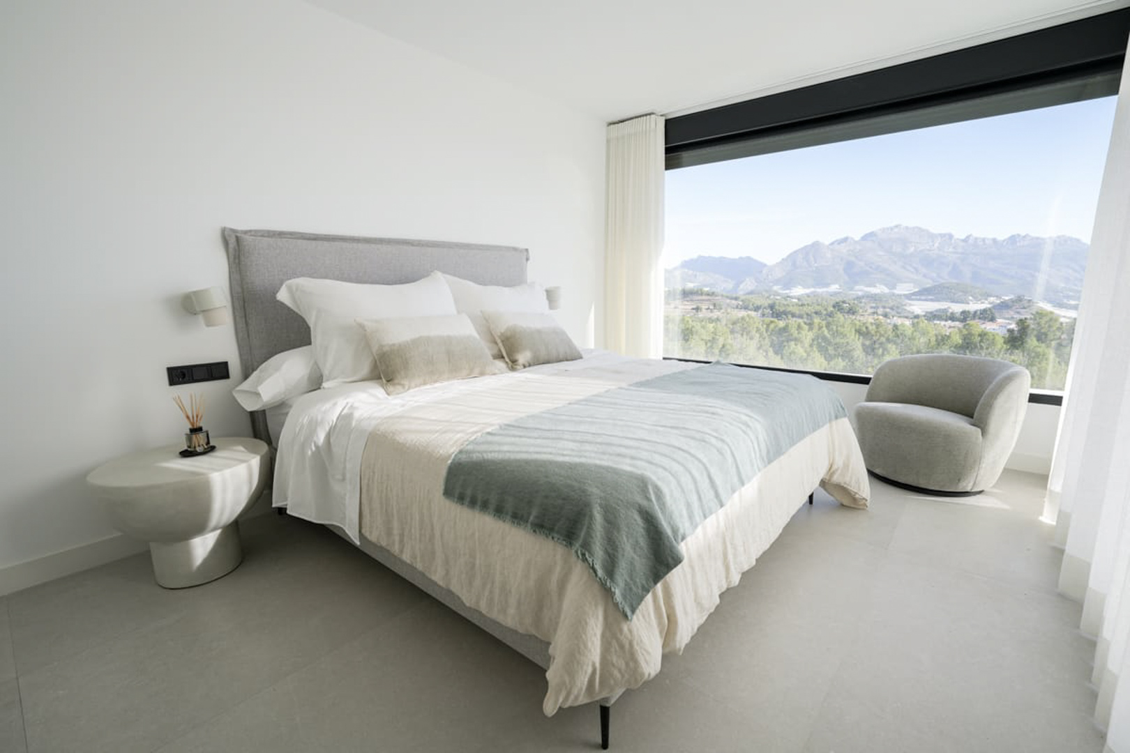Dormitorio luminoso con cama doble, butaca gris y vistas a la montaña a través de un gran ventanal.