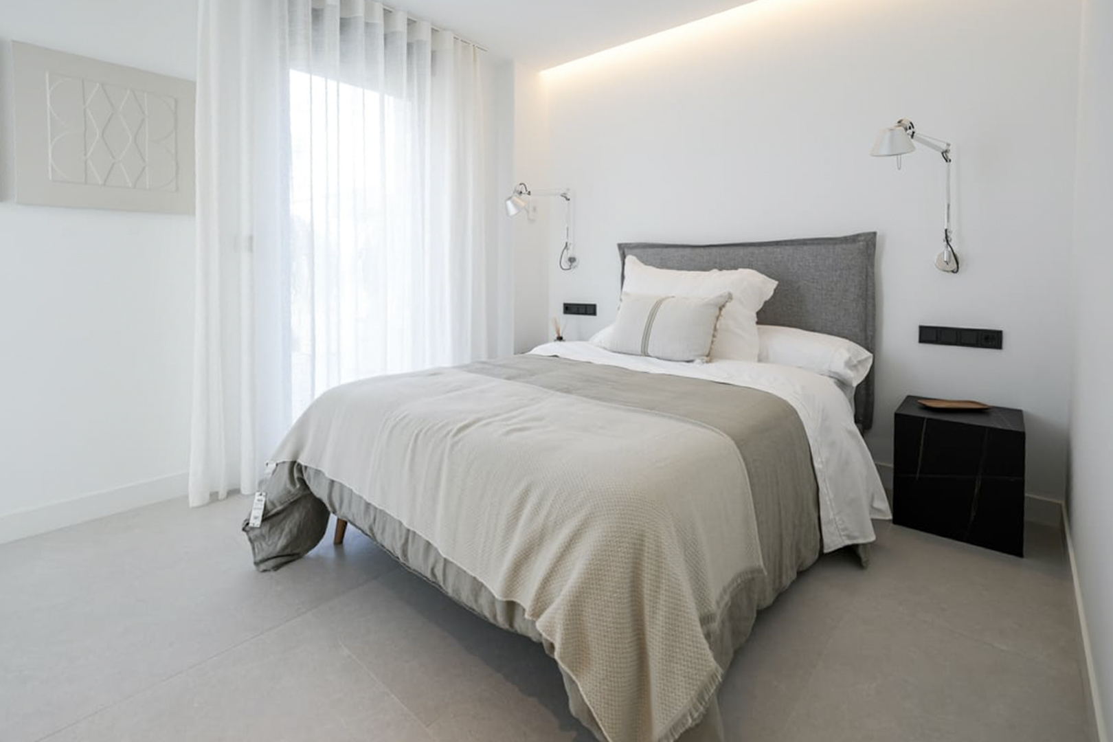 Dormitorio luminoso con cama doble, cabecero tapizado gris y lámparas de pared ajustables. Suelos de porcelanato.